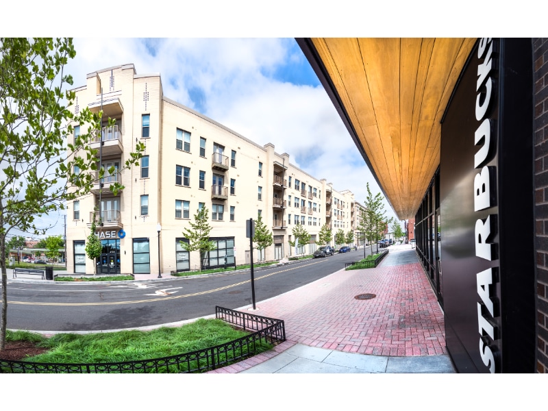 A modern apartment building with balconies near a street, featuring a Starbucks entrance visible on the right and a Chase bank on the left. Sidewalks and newly planted trees line the street, highlighting the charm of apartments in Southeast Washington DC.