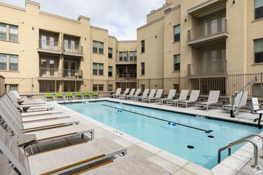 A rectangular swimming pool surrounded by lounge chairs, located in the courtyard of a multi-story apartment building. The sky is cloudy, adding a serene ambiance to the apartments at Skyland Town Center.