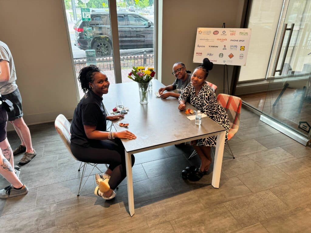 Three residents are sitting at a table near a window, smiling at the camera. A vase of flowers rests on the table, while a poster with various logos is visible in the background, showcasing our vibrant community events.