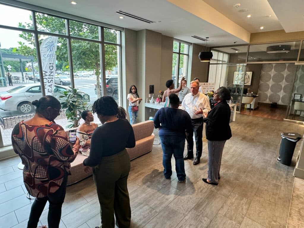 A small group of people stand and chat in a modern, open space with large windows. Some are seated, and one person appears to be checking their phone. A banner outside reads "LEASING," highlighting the vibrant community spirit and frequent resident events at Crest Apartments.