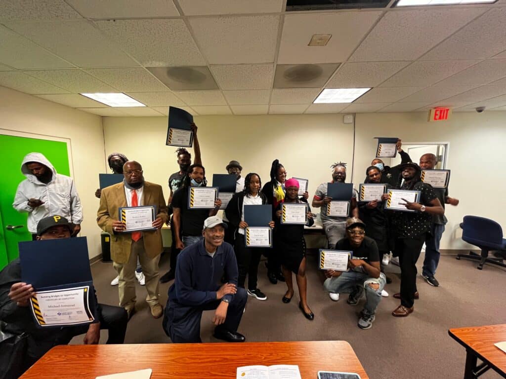 A group of people, standing and seated indoors at the Skyland Workforce Center, are holding certificates and smiling. Some are in casual clothes while others are in business attire. A green door and ceiling lights are visible in the background. It's truly a full-circle moment for everyone involved.