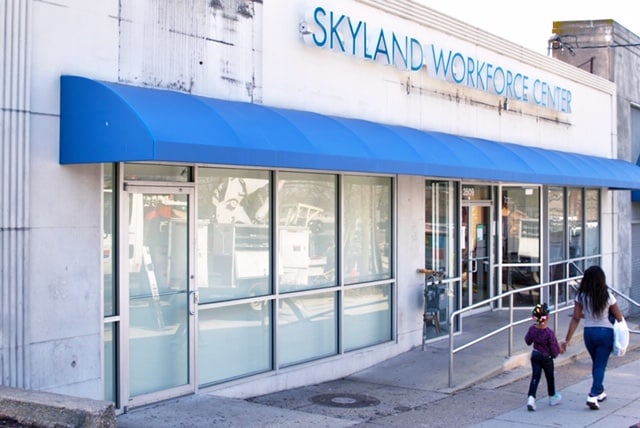 A woman and child walk toward the entrance of the Skyland Workforce Center, a beacon of hope with its blue awning and glass windows, symbolizing their journey in building the future.