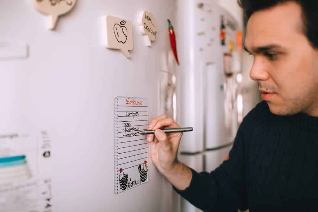 A man writes a reminder note on a refrigerator notepad to stay organized at home.