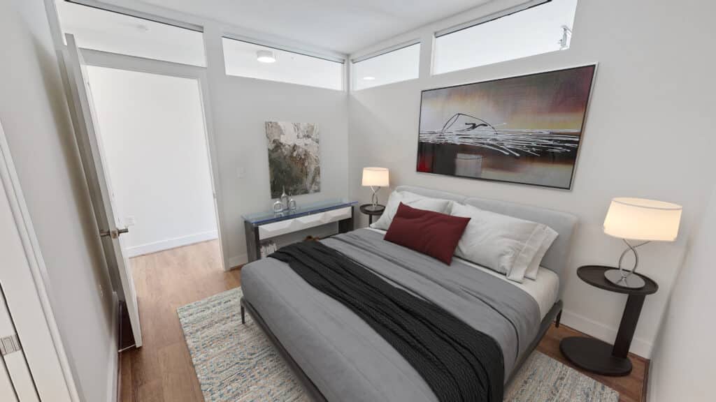 A small, modern bedroom with a double bed, gray bedding, red accent pillow, two bedside tables with lamps, a painting above the bed, a rug, and a console table with artwork.