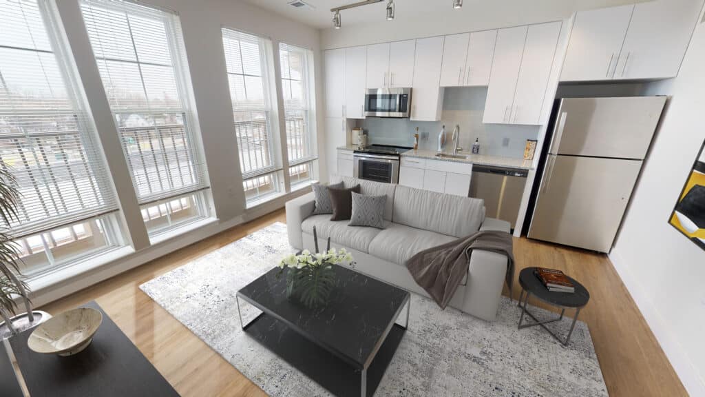 A modern living room with a white sofa, dark coffee table, and kitchen area. Large windows line the wall, and hardwood floors are visible. Stainless steel appliances are in the kitchen.