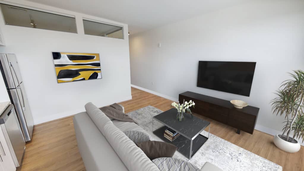 A modern living room with a gray sofa, black coffee table, large wall-mounted TV, abstract wall art, and potted plant. Wood flooring and a light wall color complete the space.