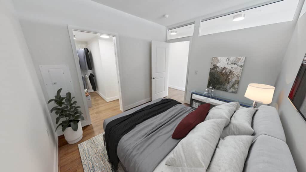 A bedroom with a neatly made bed, a bedside lamp, a plant, and an open door to a closet with hanging clothes.