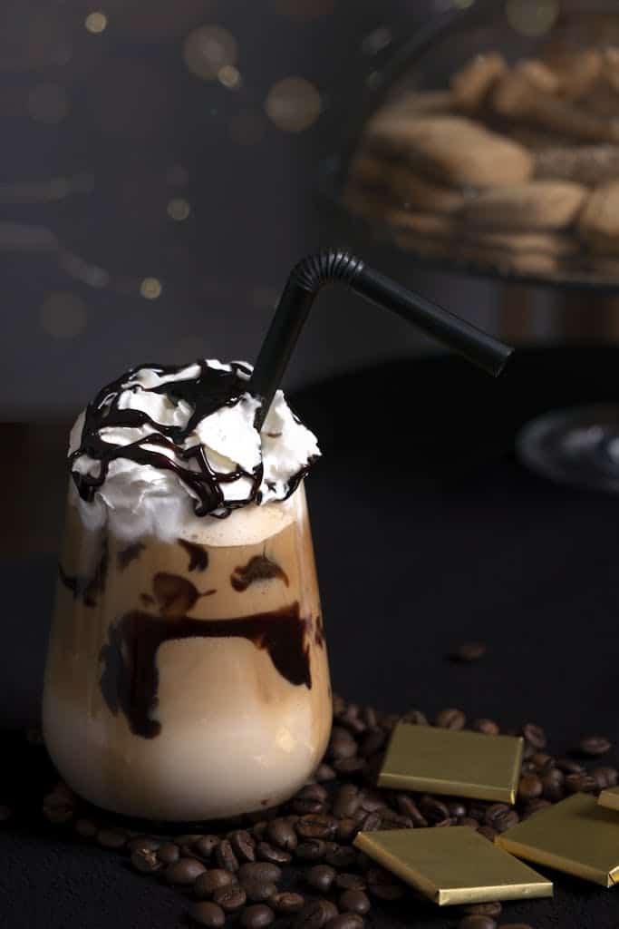 Iced coffee topped with whipped cream and chocolate syrup accompanied by coffee beans and chocolate squares.