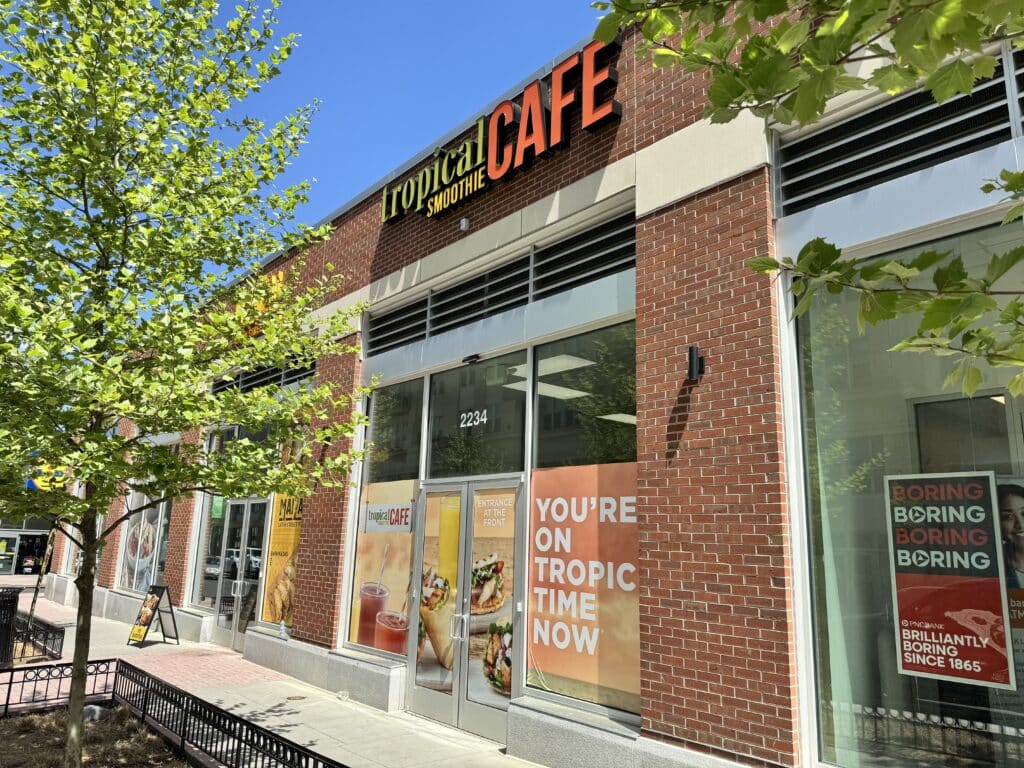 Storefront of Tropical Smoothie Cafe with promotional posters on the windows, a sidewalk, and trees in front under a clear blue sky, showcasing a vibrant lifestyle and fresh style.