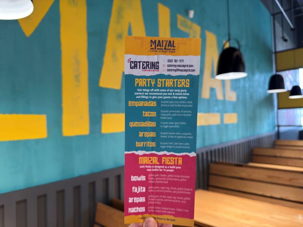 A hand holds a colorful Maizal Latin Street Food menu in front of a wall with large yellow lettering and wooden booth seating, capturing the vibrant lifestyle and style of the restaurant.